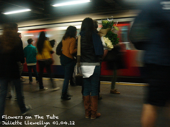 A Girl standing holdong bunches of white and green flowers on the underground platform, as a tube train arrives.